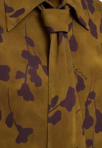 Blusa marrone con motivo floreale viola scuro, colletto a punta e dettaglio di nodo al collo; tessuto liscio con una leggera lucentezza e vestibilità sartoriale.