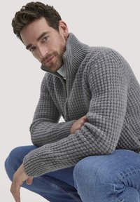 Grauer Strickpullover mit hohem Kragen und Reißverschluss, versehen mit strukturiertem Rippstrick. Kombiniert mit blauen Jeans.
