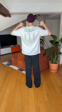 Biała koszulka z nadrukiem graficznym na plecach, noszona z czarnymi spodniami o szerokich nogawkach. Na tle naturalna drewniana podłoga i rośliny doniczkowe.