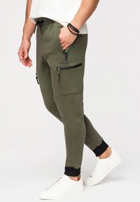 Homme portant un jogging vert olive avec des poches zippées, chevilles noires resserrées, baskets blanches et bracelets noirs à perles à un poignet.