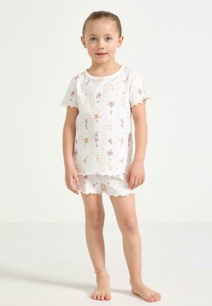 Ragazza giovane in piedi scalza che indossa un completo pigiama bianco con stampe floreali e coniglietti, maniche corte e bordi arricciati.