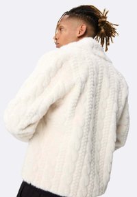 Une personne avec des dreadlocks attachées en arrière porte une épaisse veste en maille blanche texturée, légèrement tournée loin de l'appareil photo sur un fond uni.