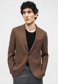 Νέος άνδρας με σγουρά μαλλιά φοράει ένα καφέ σακάκι πάνω από μαύρο πουλόβερ και γκρι παντελόνι, στέκεται με τα χέρια στις τσέπες μπροστά σε ένα απλό φόντο.