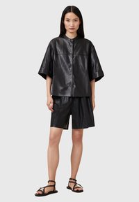 Μαύρο δερμάτινο πουκάμισο με κοντά μανίκια, κουμπιά στο μπροστινό μέρος και στρογγυλεμένο γιακά, σε συνδυασμό με σορτς στο ίδιο σχέδιο και μαύρα σανδάλια με επίπεδη σόλα. Ομαλή υφή.
