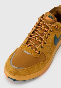 Sapatilha desportiva Nike em camurça e malha amarelo mostarda, com atacadores padrão em laranja e preto, um swoosh azul e uma sola de borracha texturizada.