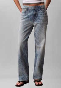 Άτομο που φοράει μπλε τζιν ψηλόμεσο με χαλαρή εφαρμογή και διακριτικό λεοπάρ σχέδιο, μαύρες σαγιονάρες, με τα χέρια στις τσέπες μπροστά σε απλό φόντο.