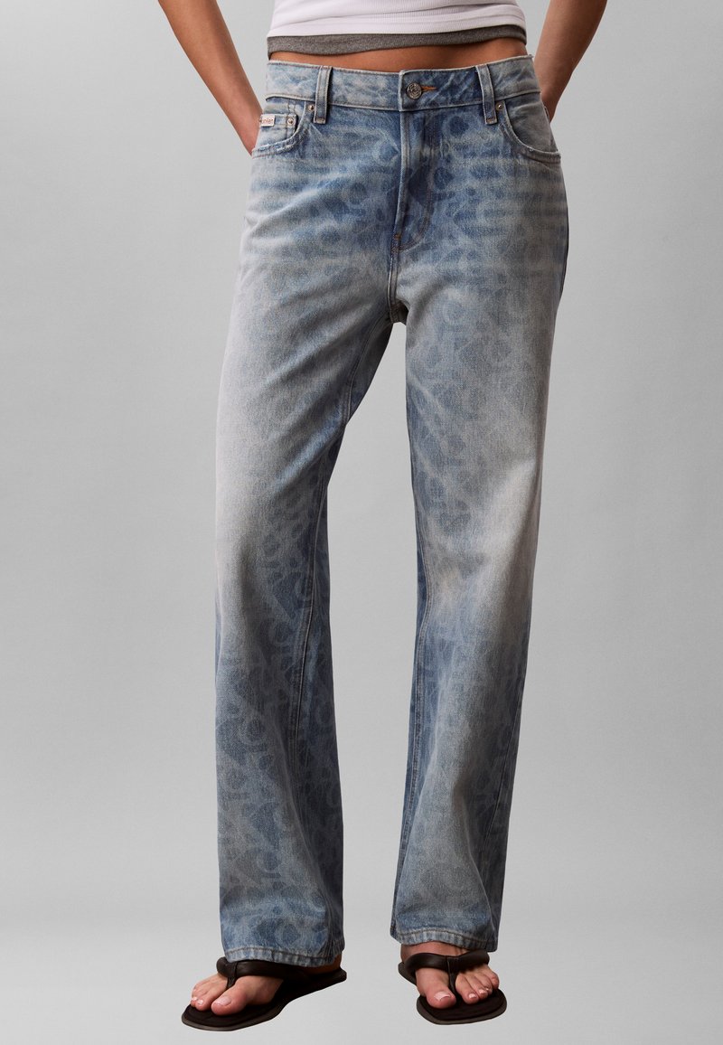 Άτομο που φοράει μπλε τζιν ψηλόμεσο με χαλαρή εφαρμογή και διακριτικό λεοπάρ σχέδιο, μαύρες σαγιονάρες, με τα χέρια στις τσέπες μπροστά σε απλό φόντο.