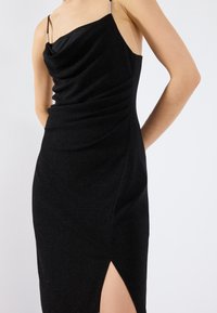 Robe noire sans manches avec un col drapé, texture plissée et une fente sur le côté. Fabriquée dans un tissu scintillant, mettant en valeur une silhouette cintrée.