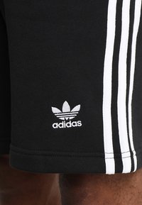 Shorts de sport noirs en tissu lisse avec trois bandes blanches le long des côtés et un petit logo Adidas blanc sur l'ourlet.