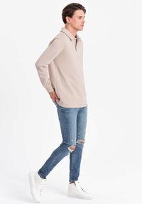Beige långärmad polo-skorta, mjukt tyg, klassisk krage, kombinerad med ljusblå rivna jeans och vita hög-top sneakers.