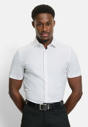 Mann trägt ein eng anliegendes weißes kurzärmeliges Hemd mit Knöpfen, schwarze Hose und schwarzen Gürtel, steht vor einem schlichten hellen Hintergrund.