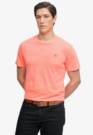 Hombre con una camiseta coral de manga corta metida en vaqueros negros con un cinturón marrón, de pie con las manos detrás de la espalda.