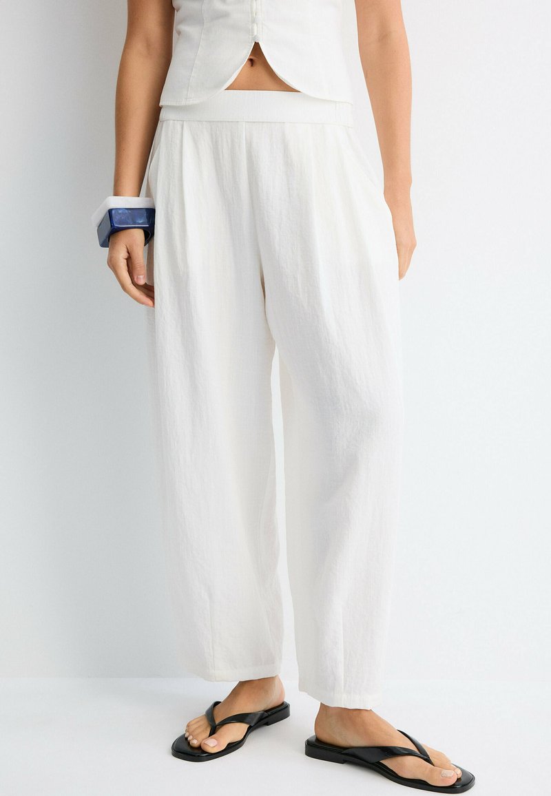 Femme portant un pantalon blanc ample, un top blanc sans manches, des sandales noires en tongs et un bracelet épais bleu et blanc au poignet gauche.