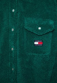 Grön fleece-tröja med en texturerad yta, prydd med silverfärgade tryckknappar och en framficka med en svart och röd "TOMMY JEANS" logotyp patch.