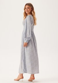 Lichtblauwe bloemenlange jurk met een gathered bodice, ballonmouwen en een vloeiende rok. Gemaakt van zachte stof, met kleine kleurrijke bloemenpatronen.