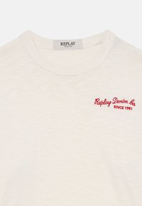 Jasno-beżowa bawełniana koszulka z okrągłym dekoltem. Na lewej piersi haftowane czerwone logo "Replay Denim Co. SINCE 1981".
