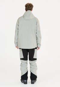 Lys grå vandtæt jakke med hætte, kombineret med sorte skibukser med grå detaljer og store sidelommer.