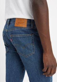 Personne portant un jean Levi's 511 slim fit bleu et une chemise blanche, montrant la poche arrière et le patch en cuir marron sur la ceinture.
