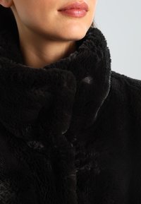 Svart fuskpälsjacka med hög krage och mjuk textur. Har en plyschfinish och sömlös design som betonar värme och komfort.