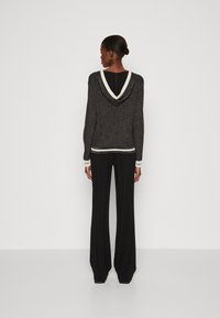 Donna con pelle scura e capelli raccolti in un chignon, che indossa un maglione con cappuccio nero con bordi bianchi e pantaloni neri larghi, di spalle contro uno sfondo semplice.