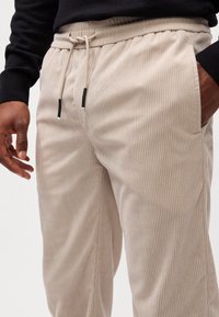 Pantalons beige côtelés avec une taille élastique et un cordon de serrage. Comprend des poches latérales et des accents noirs contrastants aux extrémités du cordon.