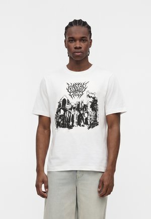 Mladý muž s vrkôčikmi, ktorý má na sebe biele tričko s čiernou grafikou gotických ruín a štýlovým textom, v kombinácii s svetlošedými nohavicami.