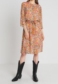 Blommig klänning med ett flerfärgat mönster i rosa, orange och grönt, med lös passform, trekvartsärmar och dragsko i midjan. Bärs med svarta cowboystövlar.