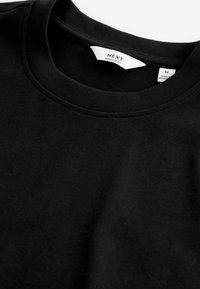 T-shirt noir col rond avec une étiquette blanche visible dans le cou indiquant la marque "NEXT" et la taille "M".