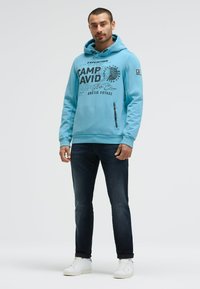 Light blue hoodie with front graphic print, adjustable drawstring, and side zippered pocket; paired with dark jeans and white sneakers.