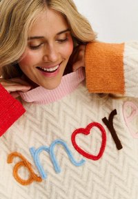 Gruby sweter w kremowym kolorze z wzorem w jodełkę. Posiada kolorowe, teksturowane haftowane litery, które tworzą słowo "amore", z kontrastującymi akcentami w kolorach czerwonym, pomarańczowym i różowym.