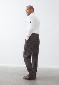 Weißer gerippter Pullover mit schwarzem Logo-Detail, braune karierte Hose und braune Leder Schuhe. Das Model posiert seitlich vor einem einfarbigen Hintergrund.