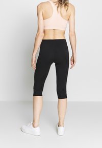 Czarne sportowe legginsy sięgające do połowy łydki o dopasowanym kroju, zestawione z jasnoróżowym sportowym stanikiem na szerokich, krzyżujących się na plecach ramiączkach oraz białymi trampkami. Gładka faktura tkaniny.