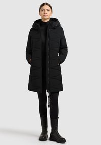 Schwarzer, gepolsterter Mantel mit Kapuze, der einen Reißverschluss vorne hat, ein geometrisches Steppmuster aufweist und einen markierten Zuglaschen-Schieber hat. Trägt er über schwarzen Leggings.