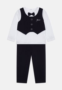 Schwarze Weste mit Knöpfen und Fliege über einem weißen Langarmshirt, kombiniert mit dunklen Leggings. Baumwollstoff, einfaches Design und verstellbarer Ausschnitt.