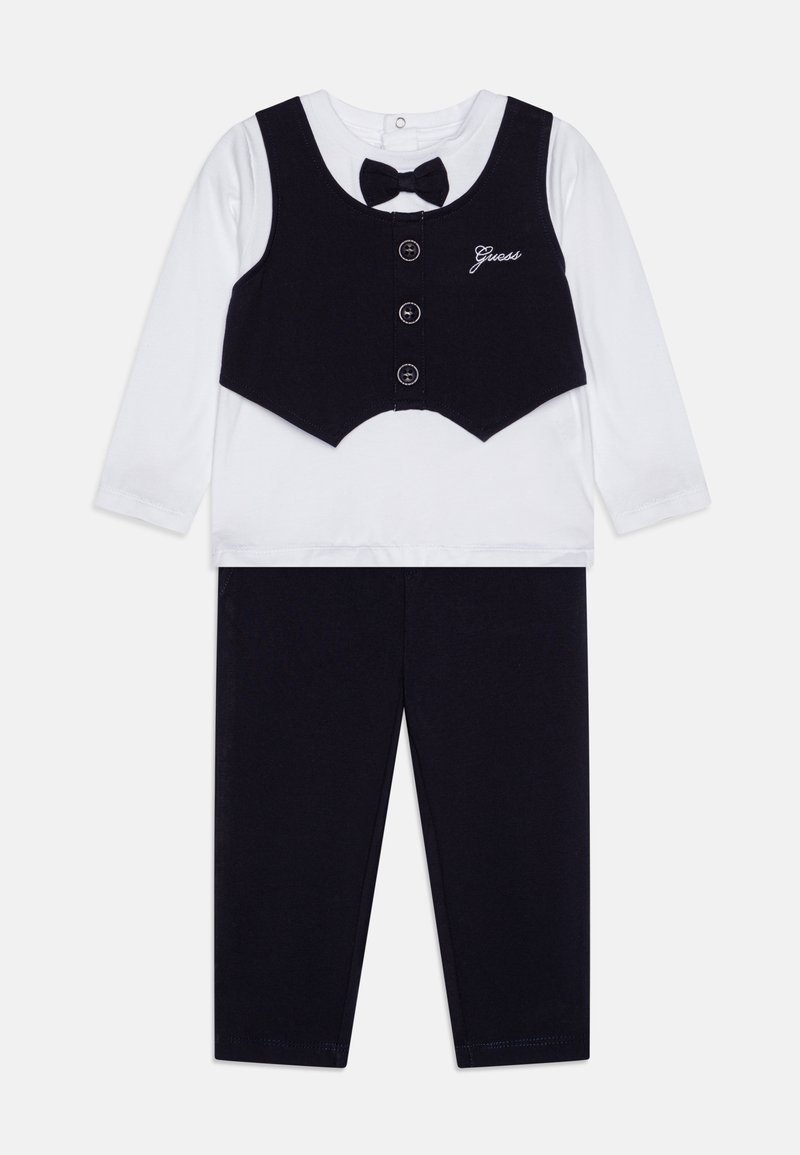 Schwarze Weste mit Knöpfen und Fliege über einem weißen Langarmshirt, kombiniert mit dunklen Leggings. Baumwollstoff, einfaches Design und verstellbarer Ausschnitt.