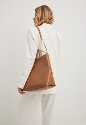 Brown leather tote bag with a smooth texture, triangular shape, and a single shoulder strap. Worn with a white blazer over loose trousers.