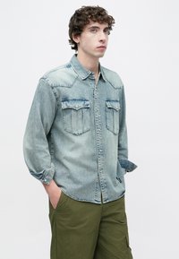 Ανοιχτό μπλε τζιν πουκάμισο με κουμπιά μπροστά, δύο τσέπες στο στήθος και μακριά μανίκια, φορεμένο πάνω από ελιάς πράσινο παντελόνι. Ομαλή υφή.