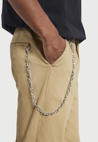 Metalen kettingaccessoire bevestigd aan beige broek, met gekoppelde segmenten en een clip. De stof is glad met een subtiele glans.