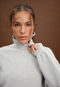 Szary, sweter z wysokim kołnierzem o teksturowanym wykończeniu. Posiada zamek błyskawiczny oraz bufiaste rękawy, noszony przez modelkę z długimi włosami.
