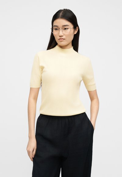 Νεαρή γυναίκα με μακριά μαύρα μαλλιά, φορώντας γυαλιά με διάφανα πλαίσια, ανοιχτόχρωμο κίτρινο κοντομάνικο τοπ και φαρδιά μαύρη παντελόνια, στέκεται μπροστά σε λευκό φόντο.