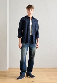 Chemise bleu marine à boutons sur un t-shirt gris, associée à un jean bleu décoloré et des chaussures sombres à semelles claires, se tenant sur un sol en bois.