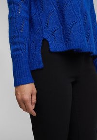 Gestrickter Pullover in lebhaftem Blau mit offenem Maschenmuster, abgerundetem Saum und Rippbündchen, kombiniert mit schwarzen, eng geschnittenen Hosen.