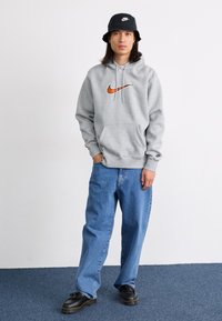 Grå hætte sweatshirt med et orange Nike-logo, parret med løse blå jeans og sorte snørebåndssko. Fladskygget sort bucket hat.