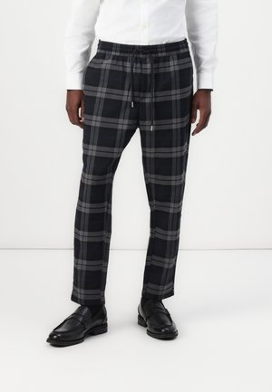 Mann trägt schwarze und graue karierte Hose mit elastischem Bund und Kordelzug, kombiniert mit schwarzen Lederloafern und weißem Langarmhemd.