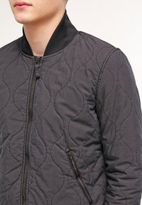 Gequilted donkergrijze bomberjack van lichtgewicht materiaal met een geribde zwarte kraag en een ritszak aan de zijkant.