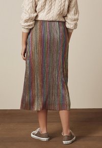 Mujer vistiendo una falda midi a rayas metálicas y multicolores, un suéter de punto trenzado beige y zapatillas Converse marrones, de pie sobre un suelo de madera.