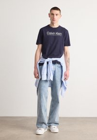 Marineblauwe Calvin Klein t-shirt met wit logo, lichtblauwe button-up overhemd geknoopt om de taille, vervaagde blauwe spijkerbroek en witte sneakers.