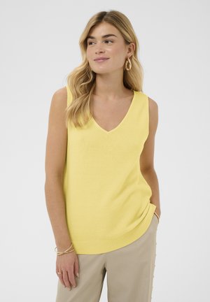 Blonde Frau trägt ein gelbes ärmelloses V-Ausschnitt-Oberteil und beigefarbene Hose, lächelt und schaut nach links, dabei eine Hand in der Tasche.