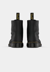 Een paar zwarte leren enkellaarzen met gele stiksels en trekflappen met de tekst "MET BOUNCING SOLES," van achteren weergegeven op een witte achtergrond.