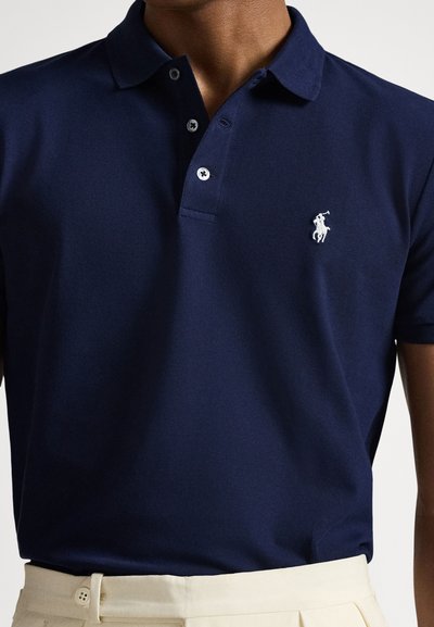 Μπλε σκούρο πόλο μπλουζάκι από υφασμάτινη υφή, με πλακέτα με τρία κουμπιά και κεντημένο λευκό λογότυπο στο αριστερό στήθος.